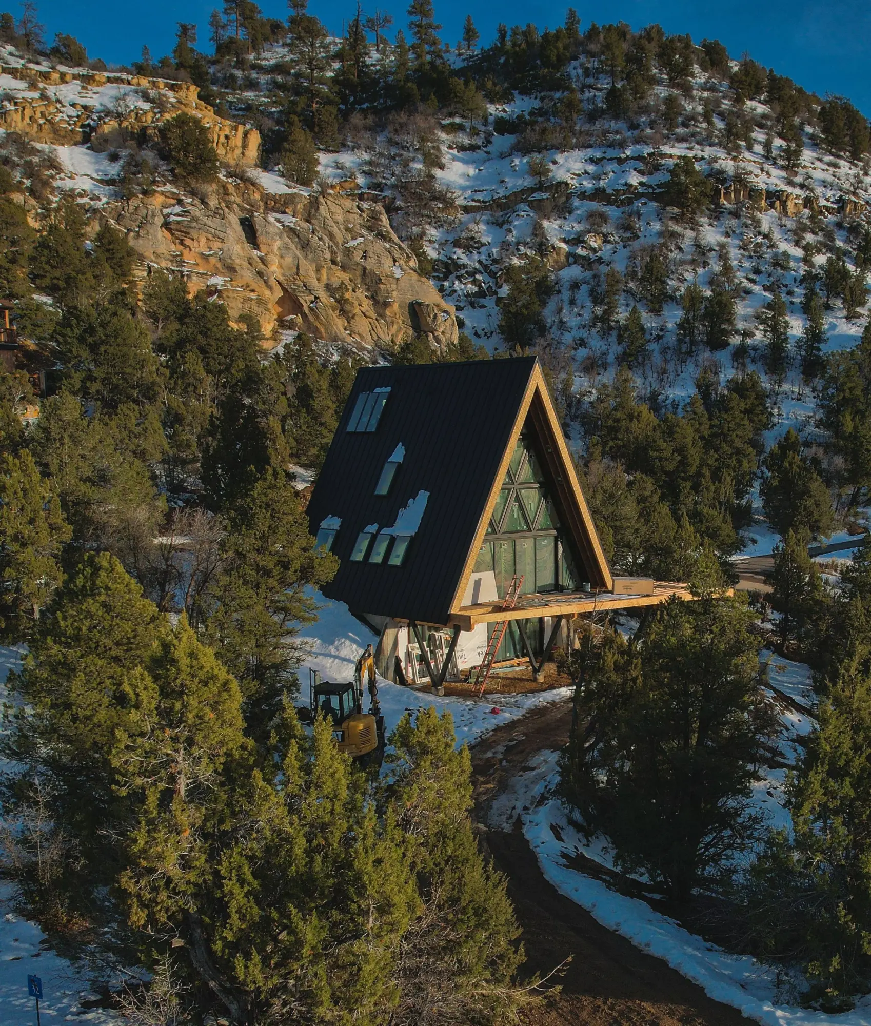 Arka during the build: the steep black roof finished on a snow-covered hillside among evergreens, a rocky mountain behind under a clear blue sky.