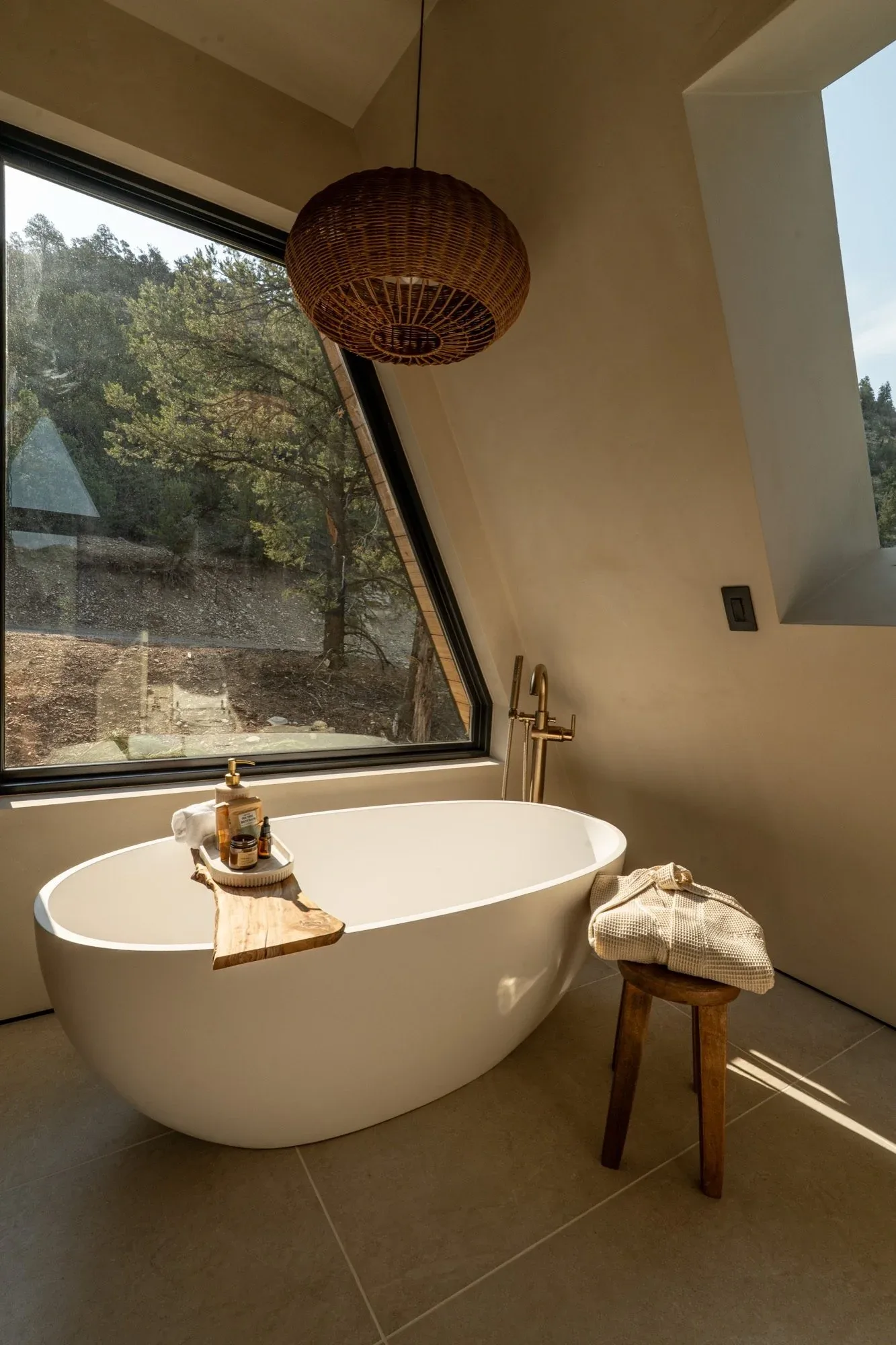 A freestanding soaking tub beside a large angled window looking out at the trees, a wooden tray of bath things across it.