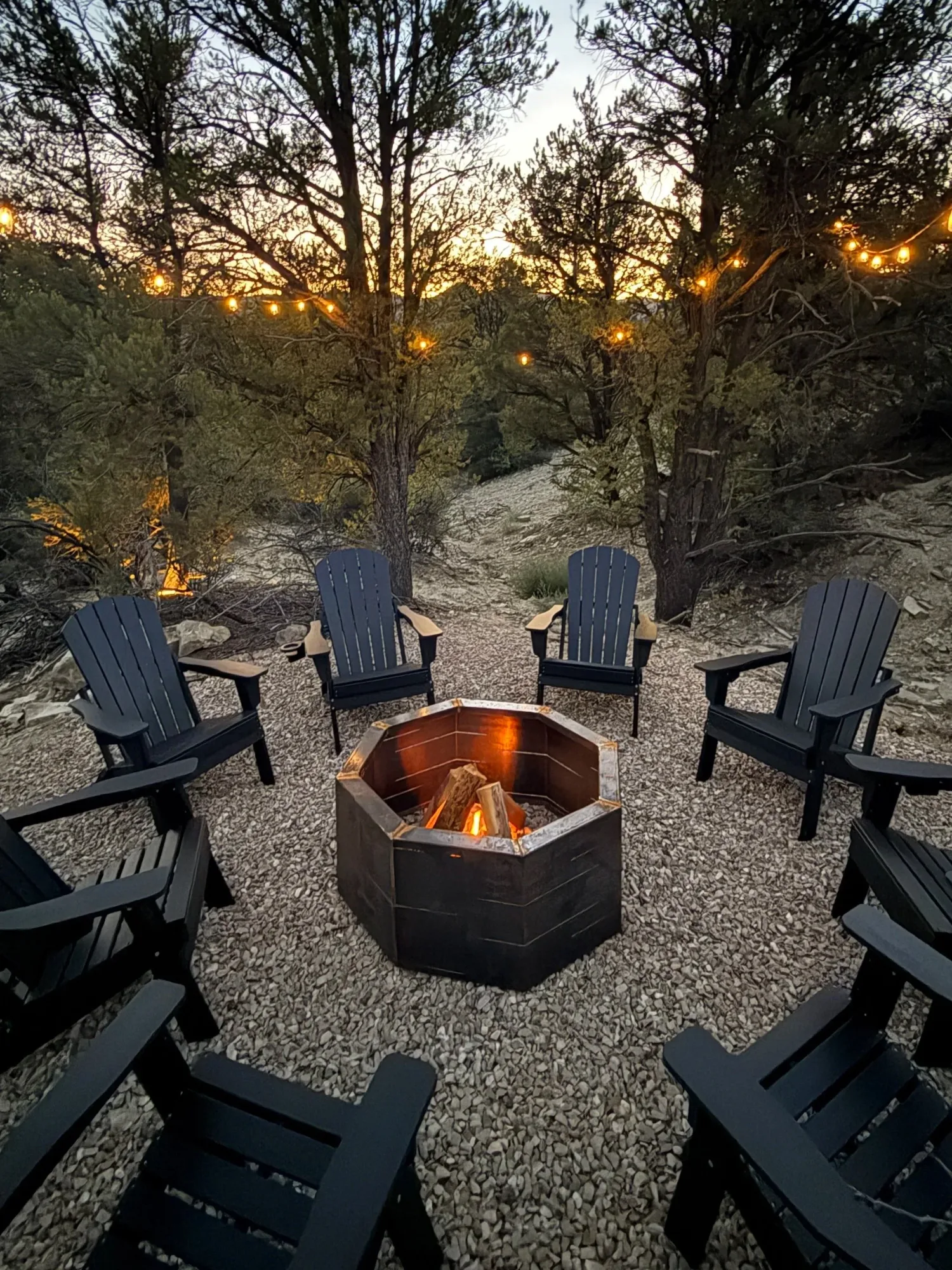 Chairs around a lit firepit on a gravel terrace among the trees at dusk.