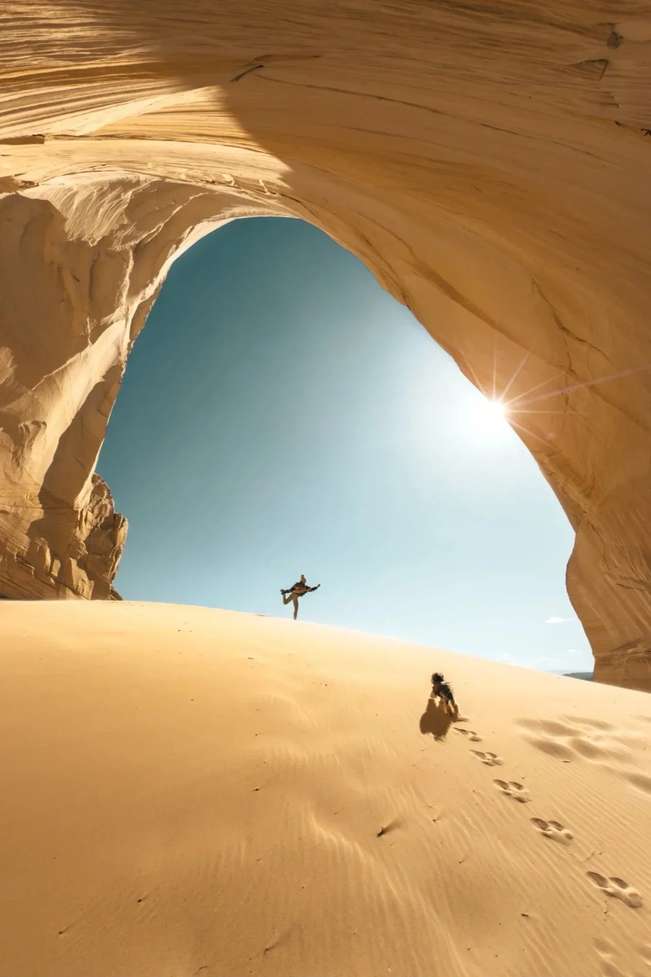 A person leaps on the sand inside the Great Chamber, a huge sandstone alcove opening to the sky and the sun.