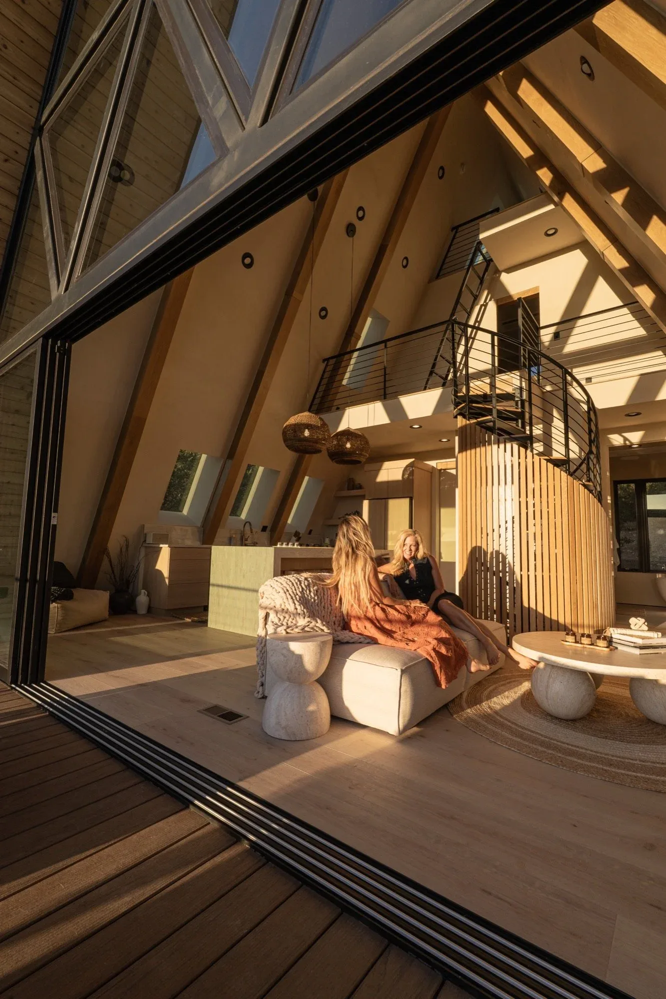 Two women talk on a white sofa in the sunlit great room, under sloped wooden ceilings and tall windows.
