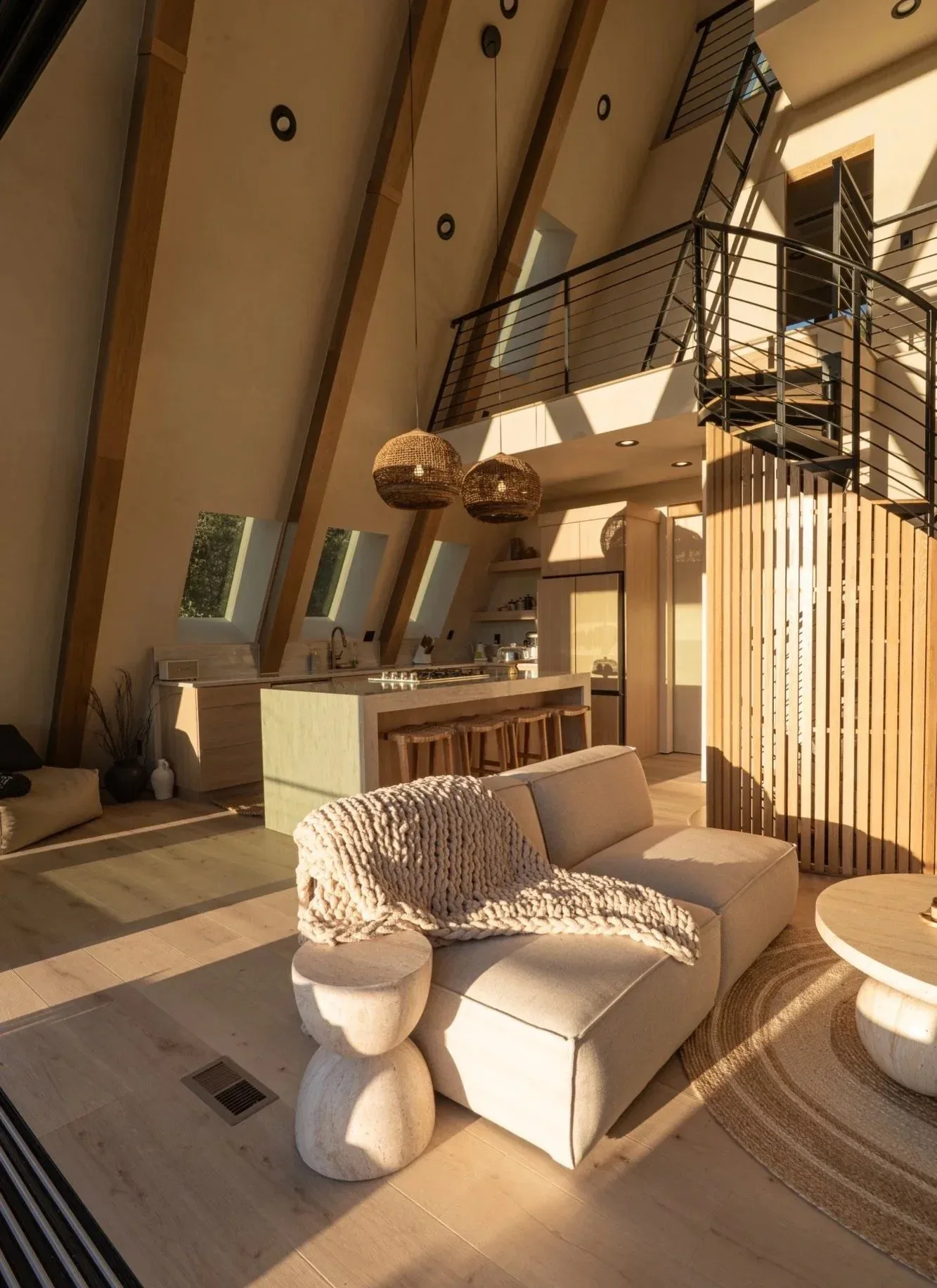 The great room: sloped wooden ceilings, tall windows, a sofa with a chunky knit throw, and a wooden kitchen island with barstools under hanging pendant lights.