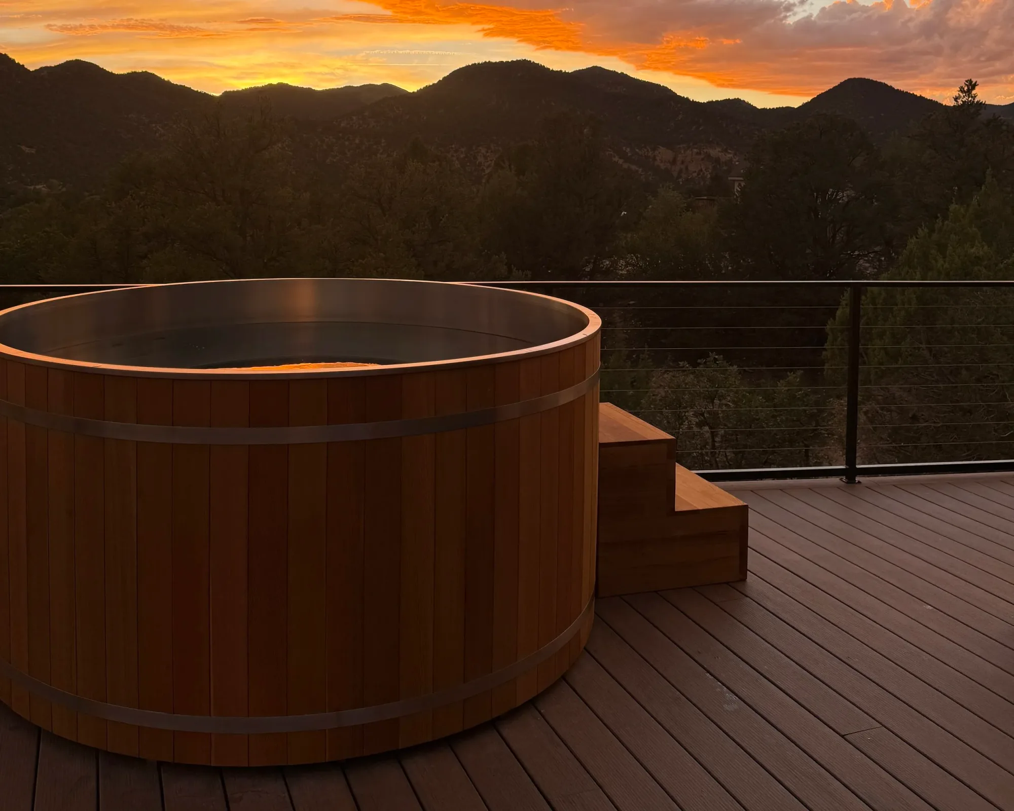 A round cedar hot tub on the deck, the ridgeline glowing orange at sunset beyond it.