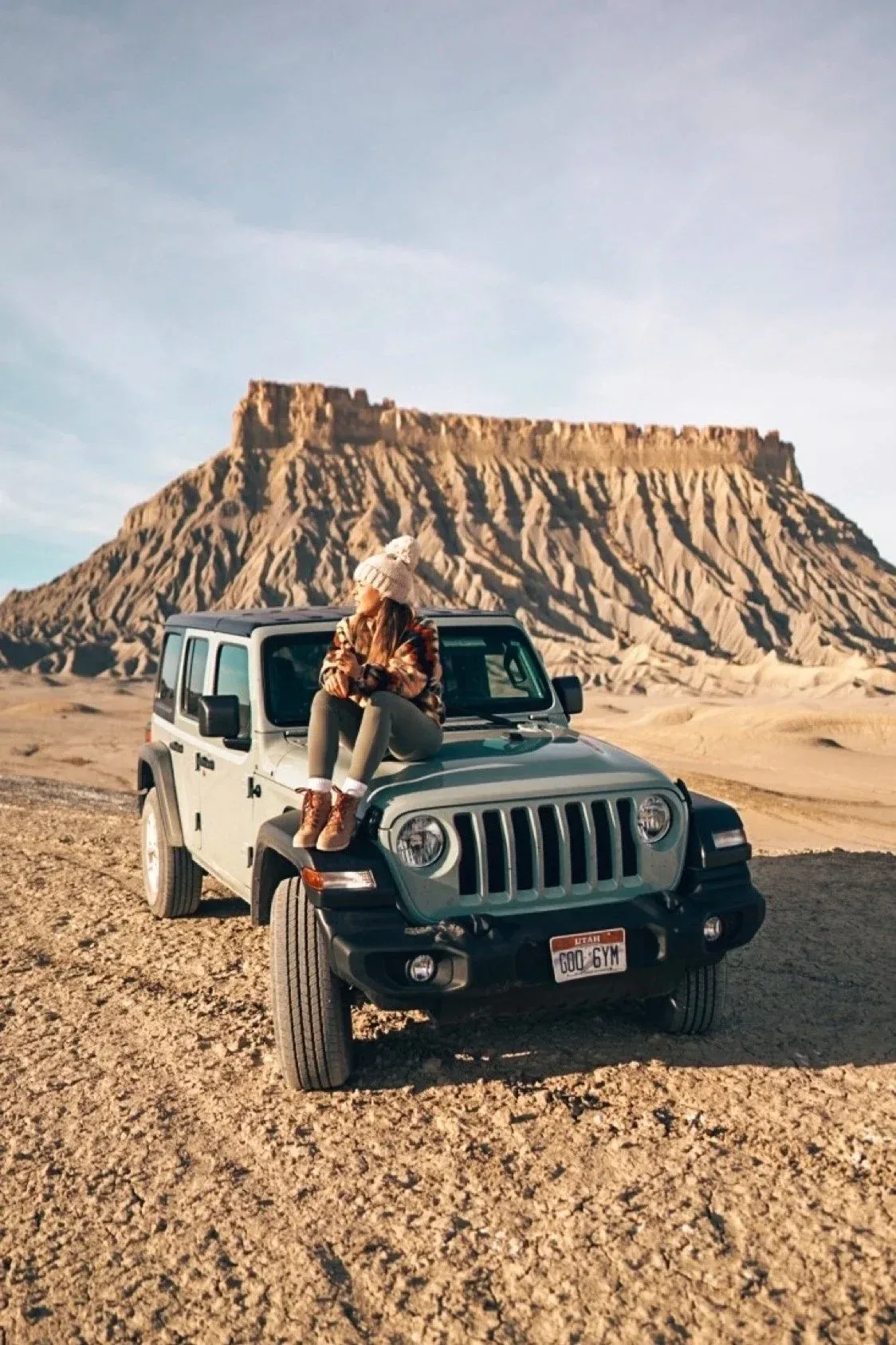 A woman sits on the hood of a Jeep in the desert, a large flat-topped mesa behind her at sunset.