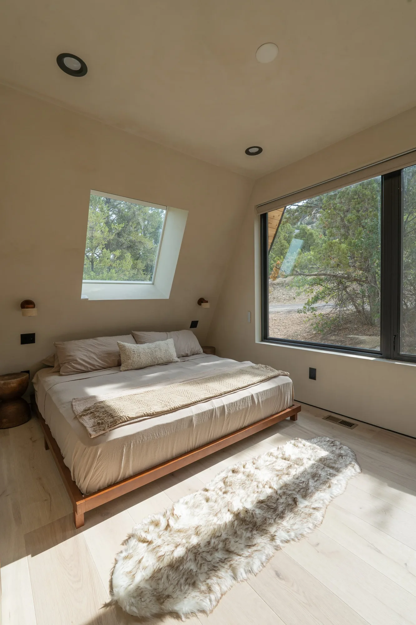 A king bedroom with white bedding, sun falling across the floor from a tall window onto the trees outside.