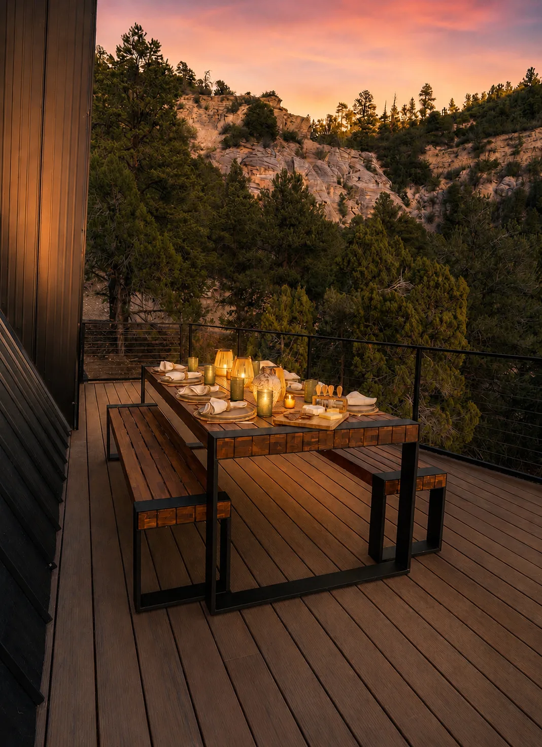 The outdoor dining table set with candles and dishes on the wooden deck at sunset, looking over forested hills.