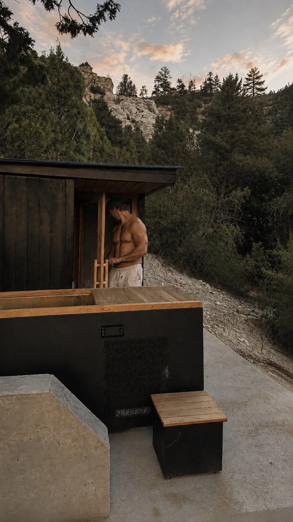 A man in a cap stands beside the wooden sauna and cold plunge outdoors, among trees and landscape rocks at sunset.