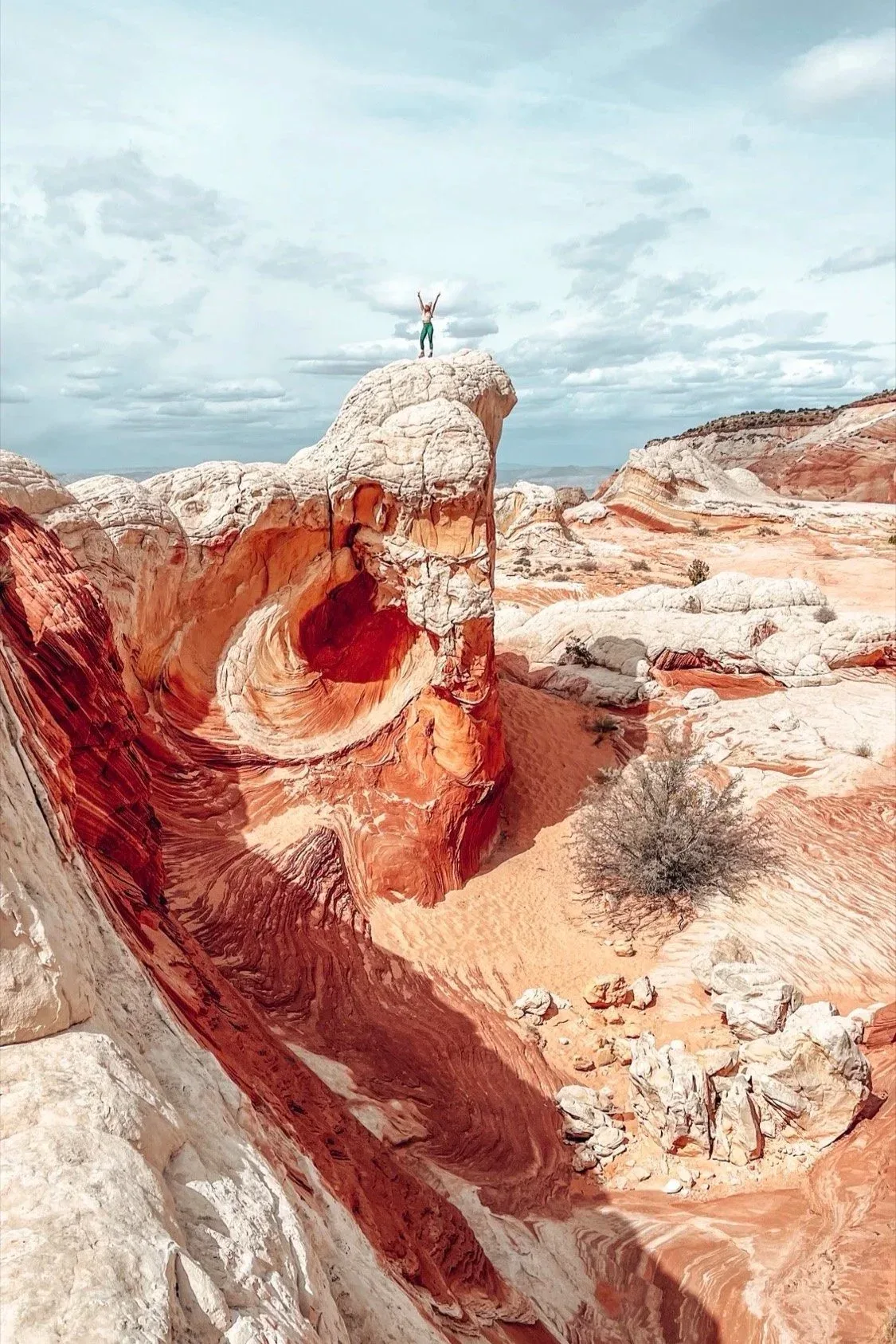 A person stands on top of swirling red, white and orange sandstone at White Pocket under a partly cloudy sky.